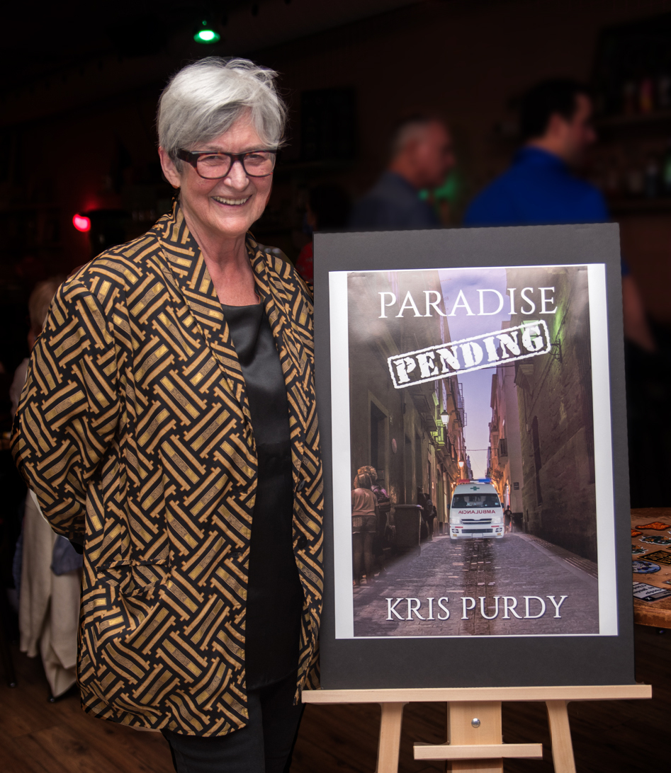 Author Kris Purdy stands next to an easel showing the cover of her book, Paradise Pending